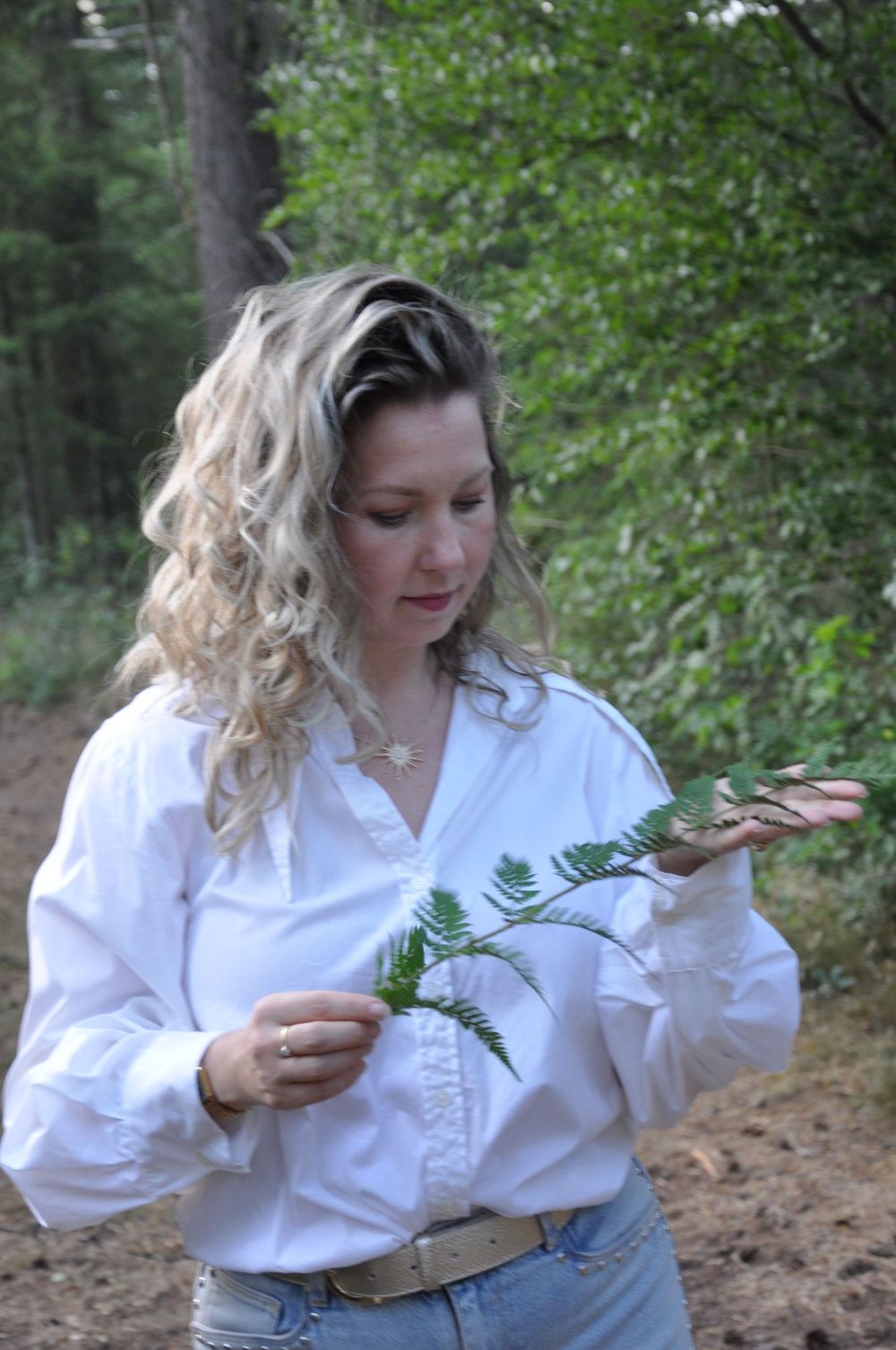 Woman with curly blonde hair in white blouse examining fern leaves in a forest setting.