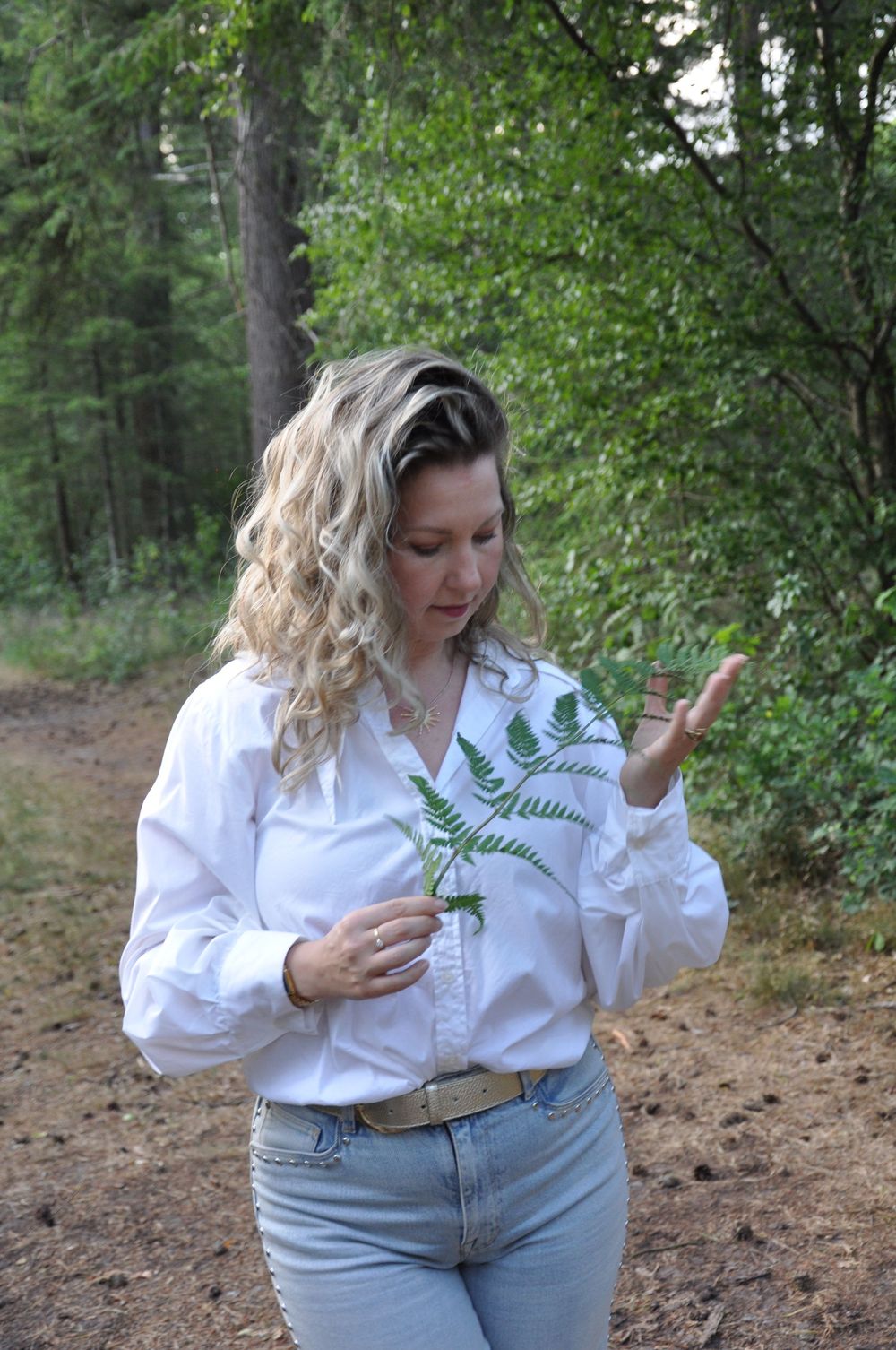 Curly blonde woman in white shirt examining a green fern frond on a forest path.