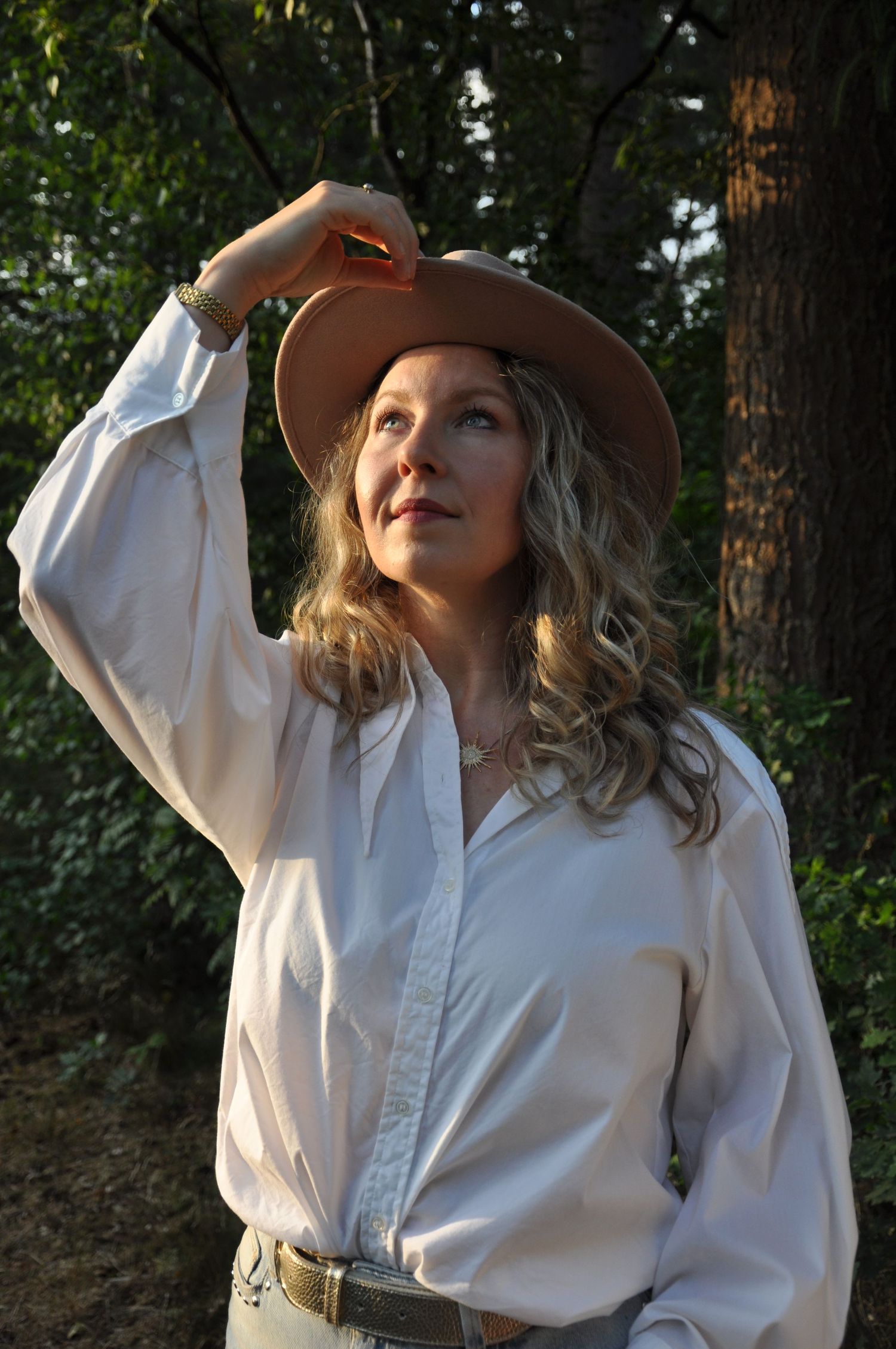 Blonde curly-haired woman in white blouse wearing brown cowboy hat looking up in sunlit forest.