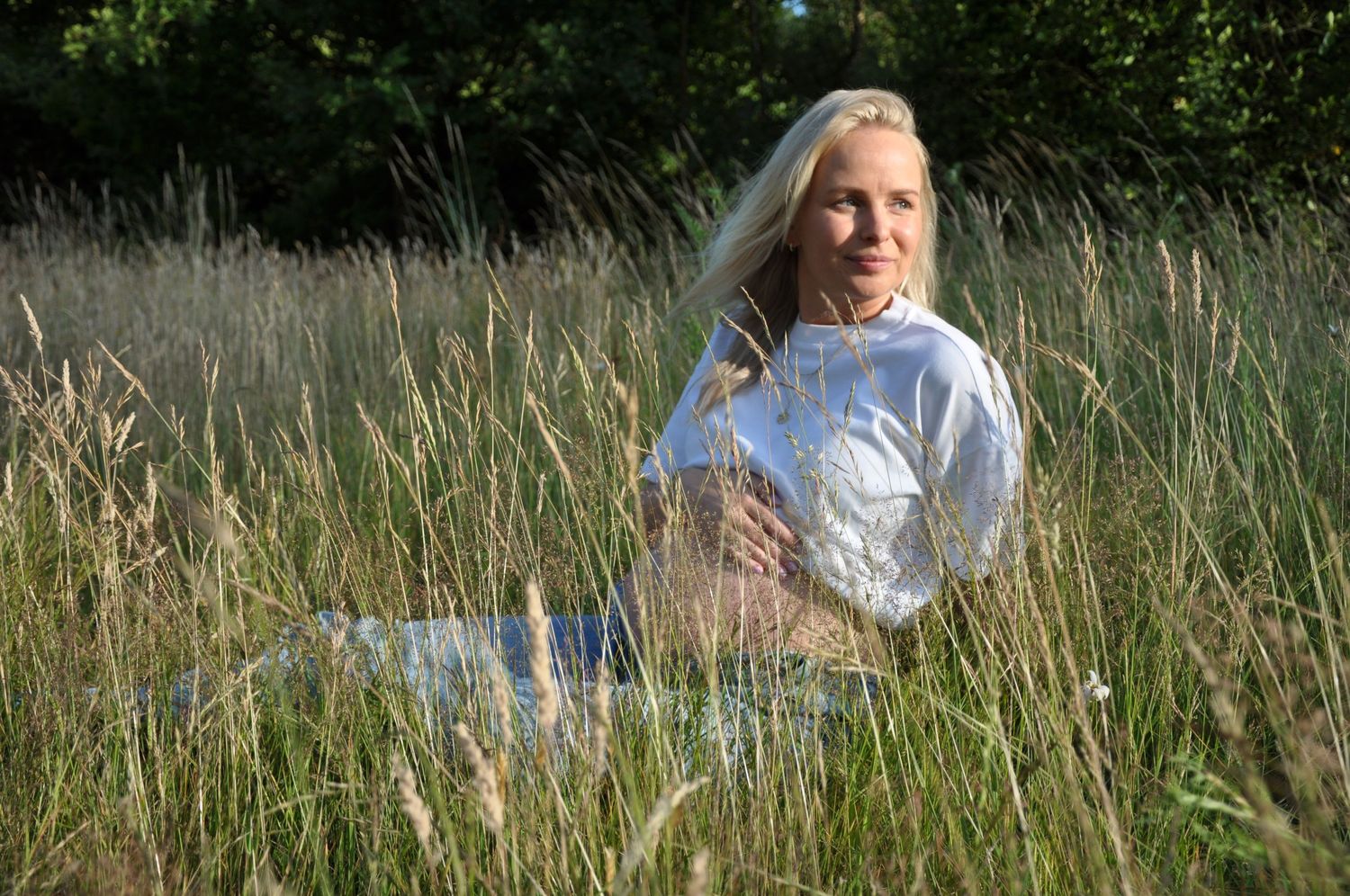 Pregnant blonde woman sitting peacefully in a sunlit meadow with tall golden grass and green trees in background.