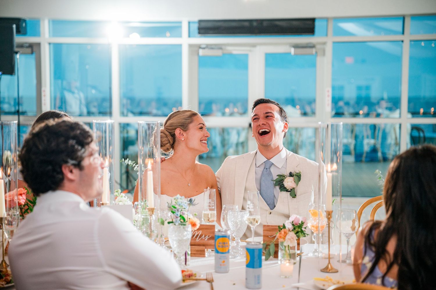 Wedding guests laugh and enjoy themselves during a reception in a bright, sunlit venue with ocean views.