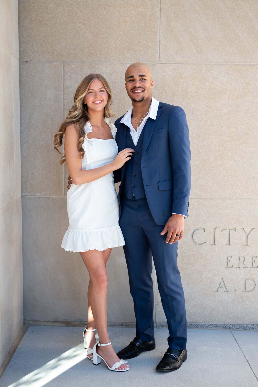 A couple in formal attire poses together against a stone wall on their wedding day.