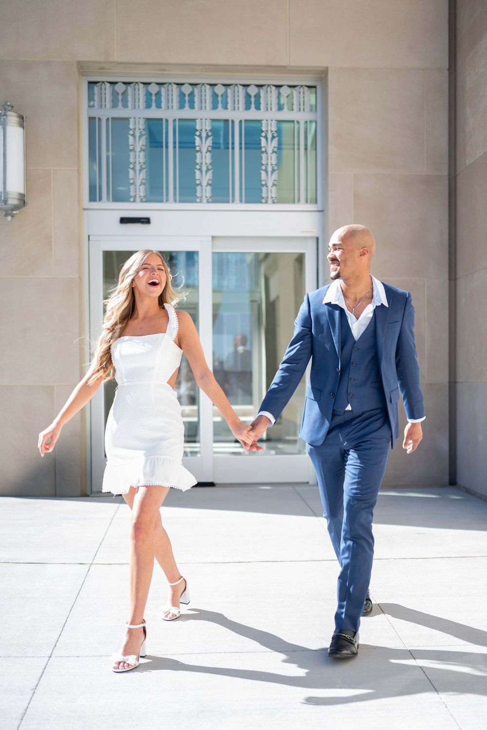 A couple in formal attire playfully walks through a sunlit corridor with dramatic shadows.