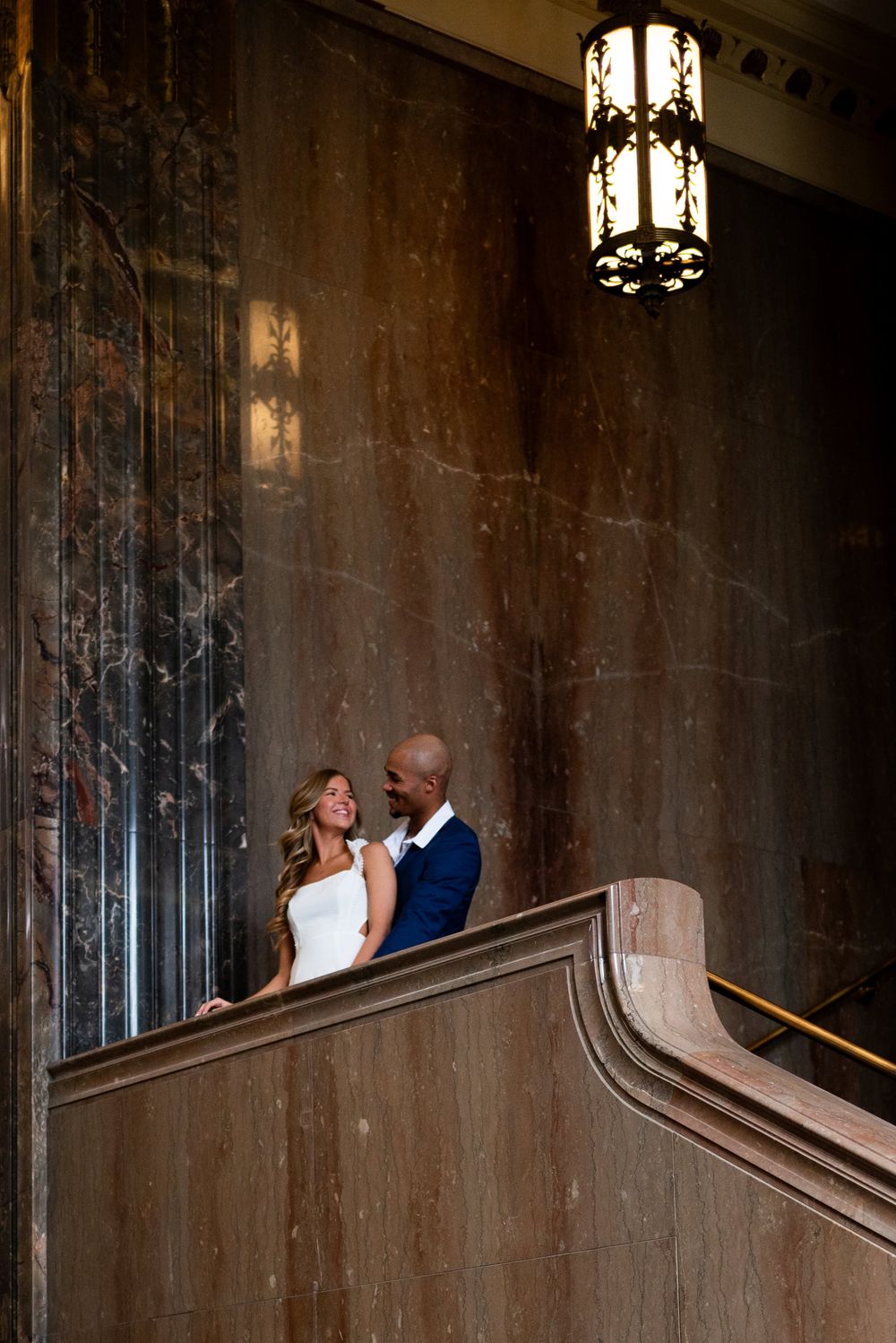 Romantic couple poses along ornate vintage staircase railing in moody lighting.