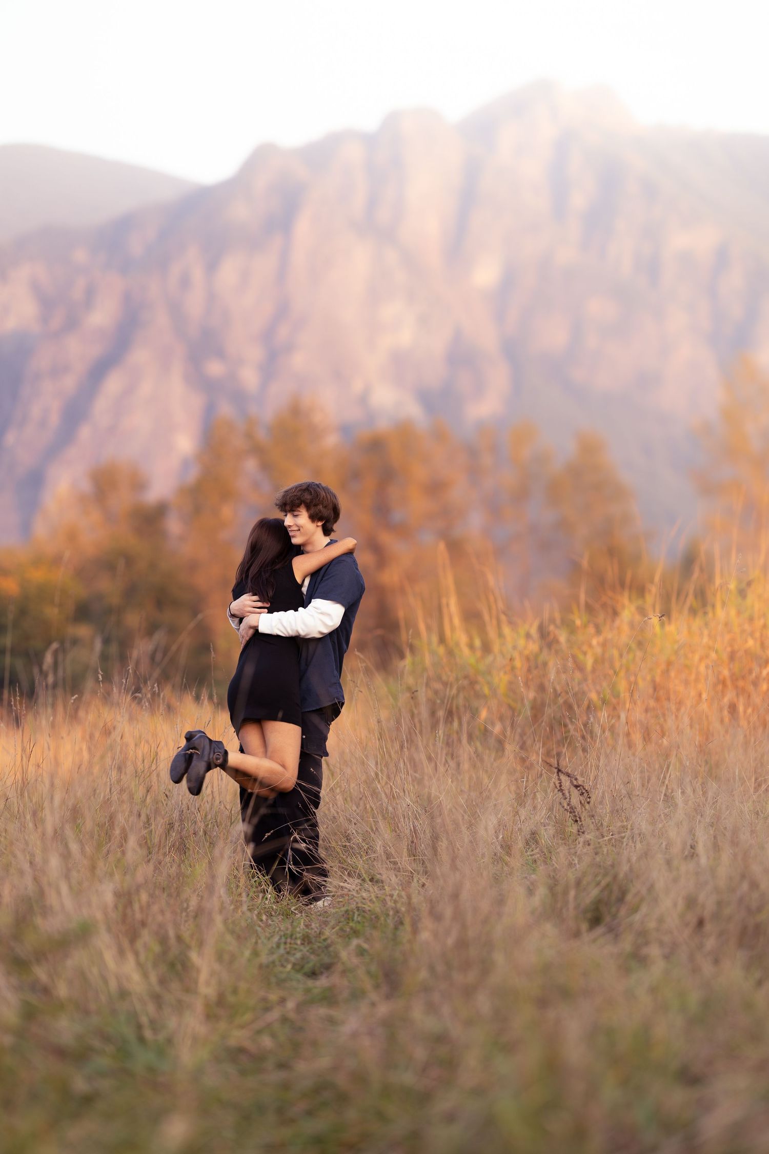 A couple embraces in a golden field at sunset with majestic mountains creating a dramatic backdrop in warm autumn light.