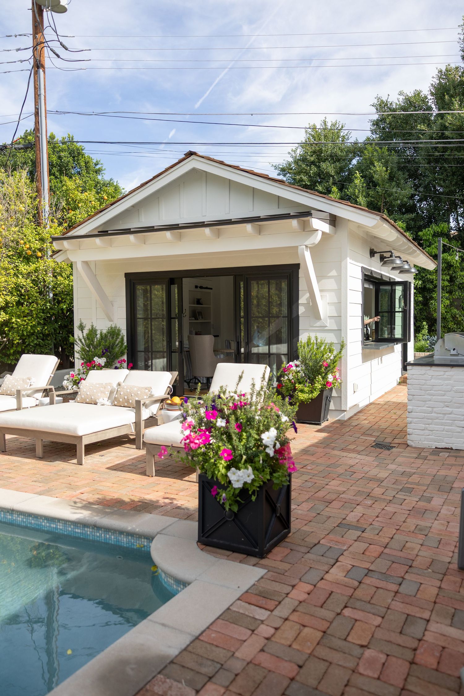 Modern white pool house with brick patio and swimming pool featuring pink flowers in planters. Arcadia Pool House Jen Wilbur Photos