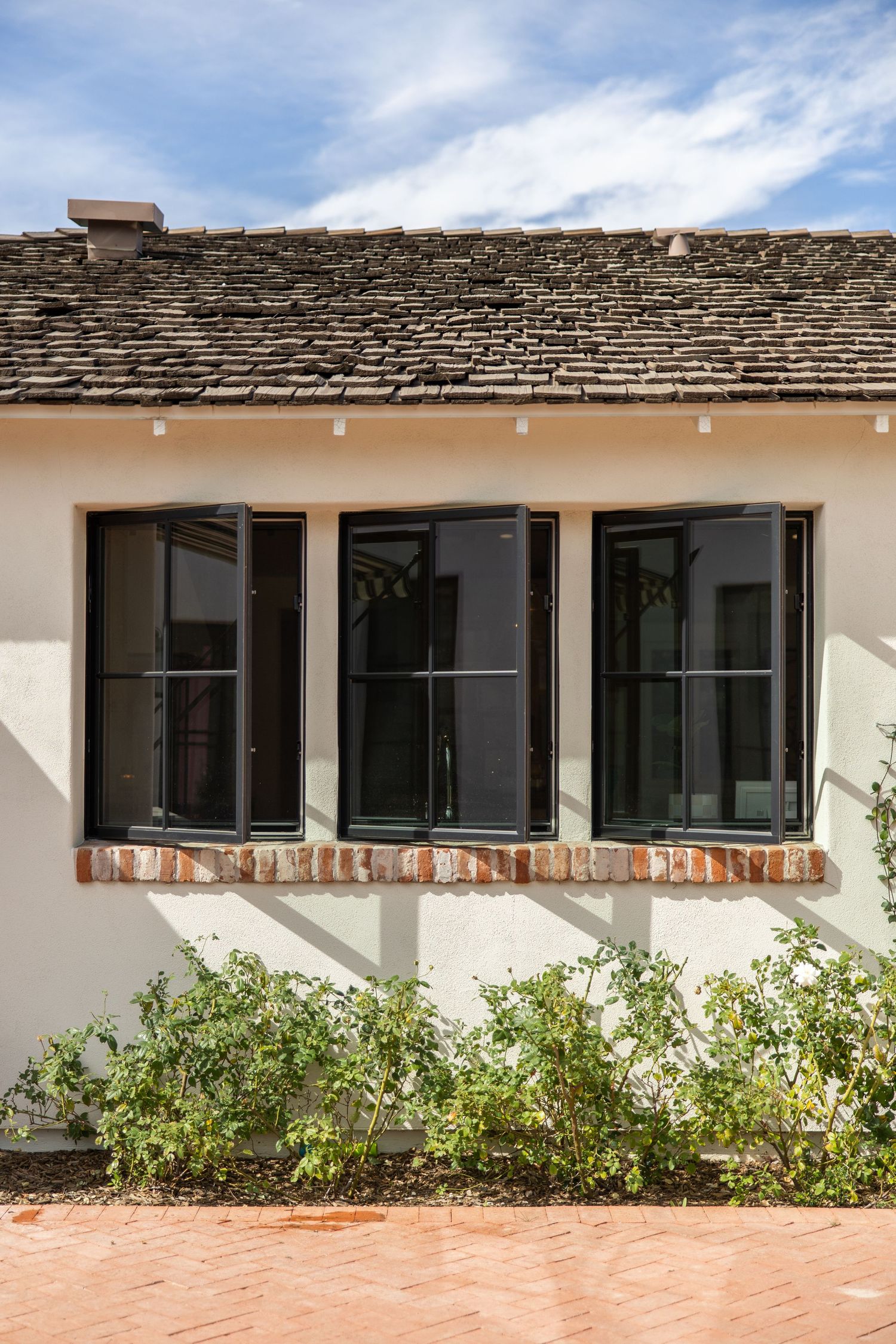 Row of dark-framed windows on white stucco wall with stone roof tiles and green shrubs below.
