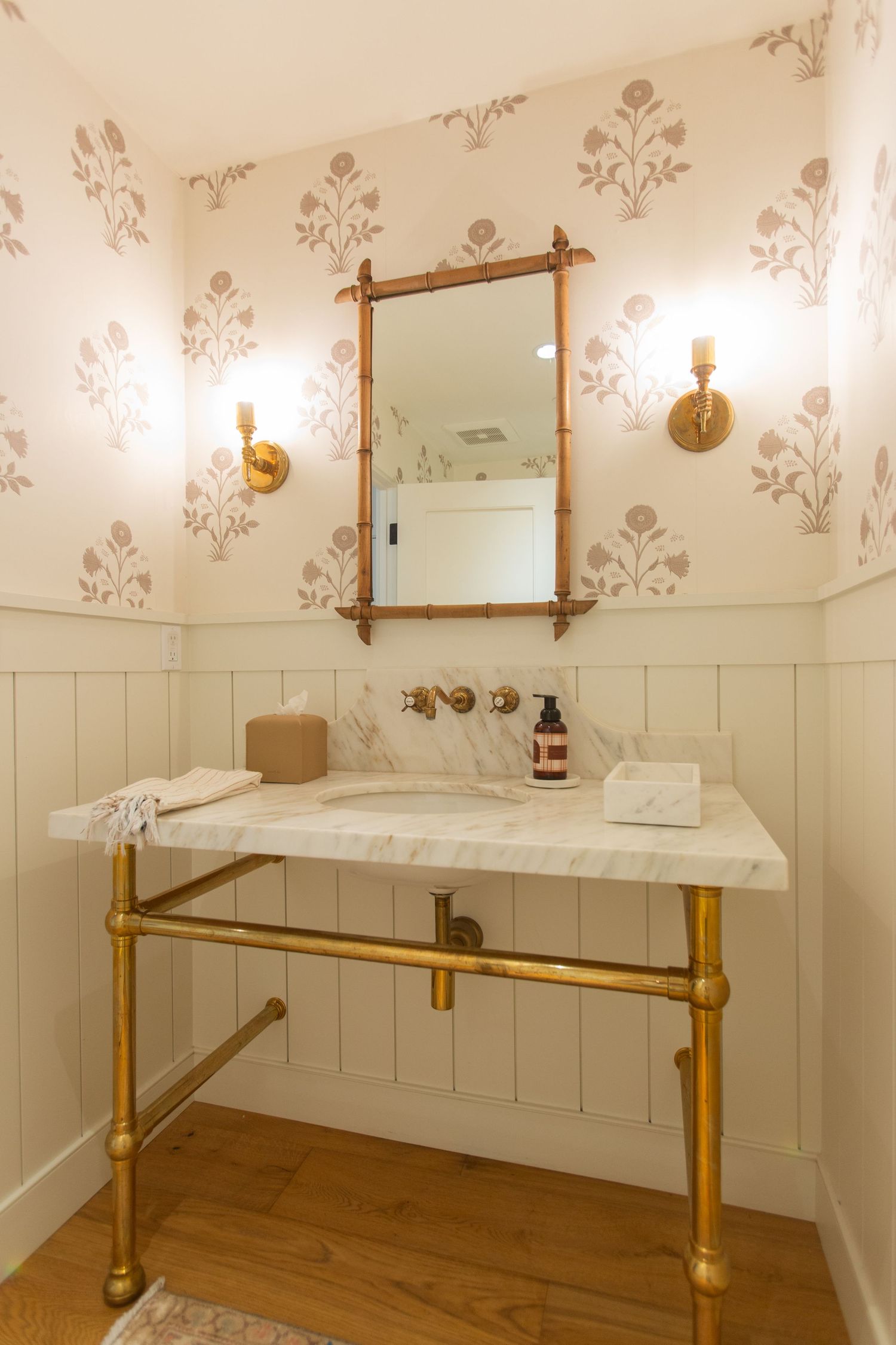 Elegant bathroom vanity with brass fixtures, marble countertop, and floral wallpaper featuring a bamboo-framed mirror. Jen Wilbur Photos