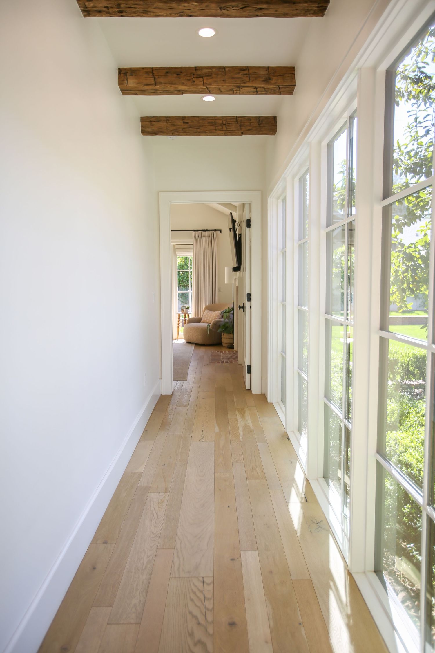 Modern hallway with white walls, exposed wooden beams, and light hardwood floors illuminated by natural sunlight. Jen Wilbur Photos, Scottsd