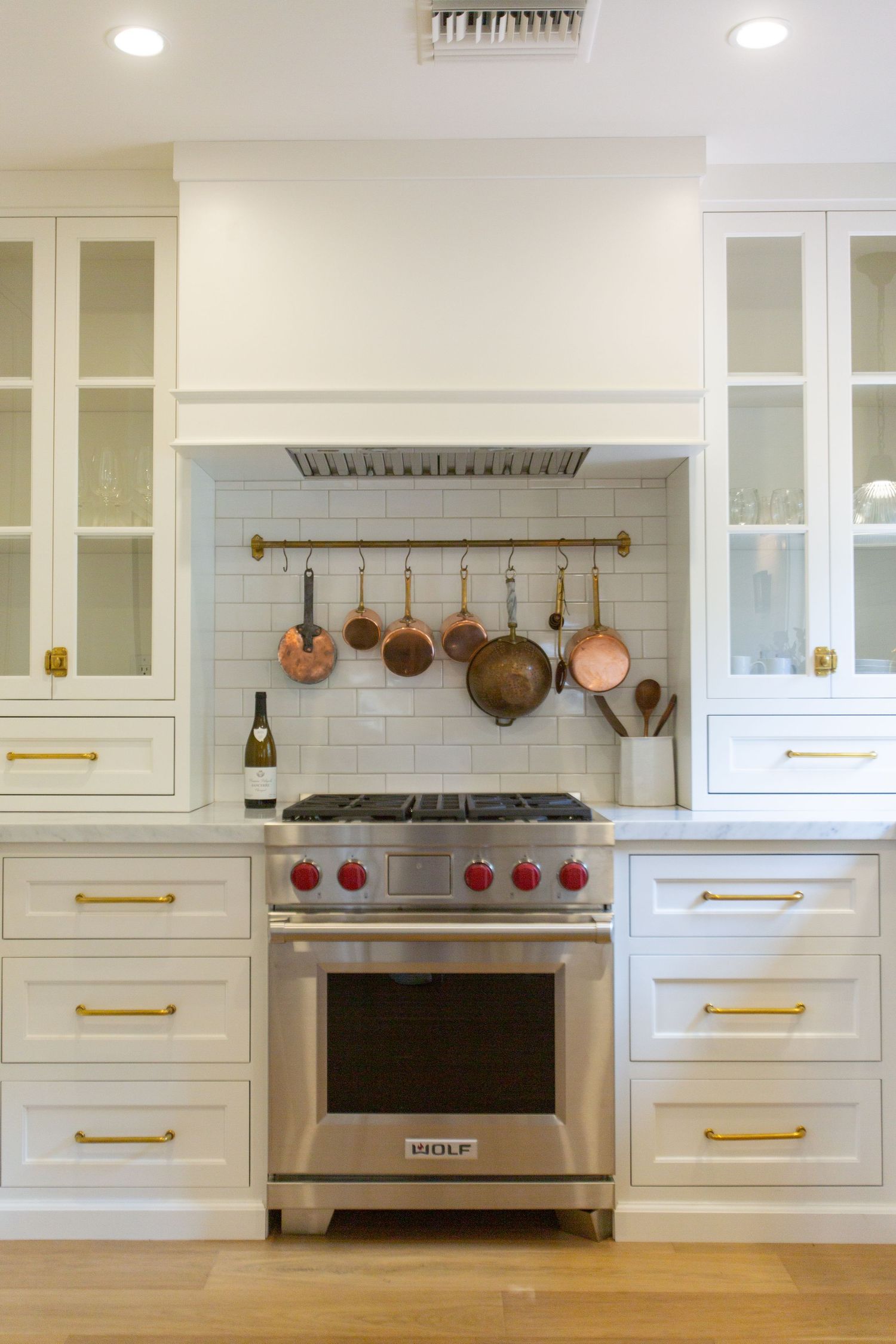 Modern white kitchen with brass hardware features a professional range and copper pots displayed on a wall-mounted rack. Jen Wilbur Photos