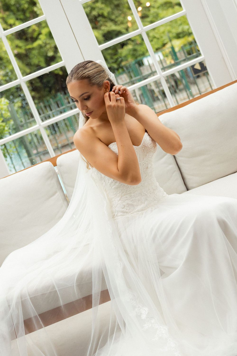 Bride in white lace dress putting on earrings while sitting in sunlit room. Wedding Photographer and videographer. Bridgewater estate