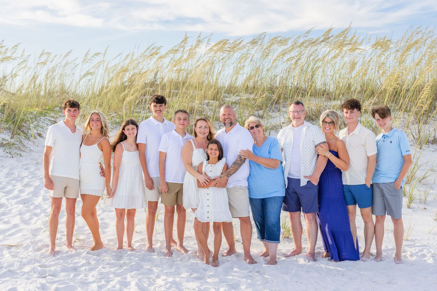 A group in coordinating white and blue outfits poses together on a sandy beach with sea grass swaying in the background.