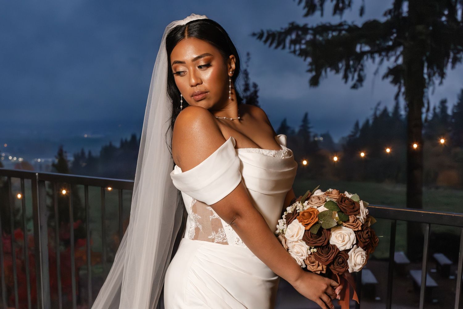 A bride in an elegant white wedding dress holds a bouquet during an evening outdoor wedding reception with string lights.