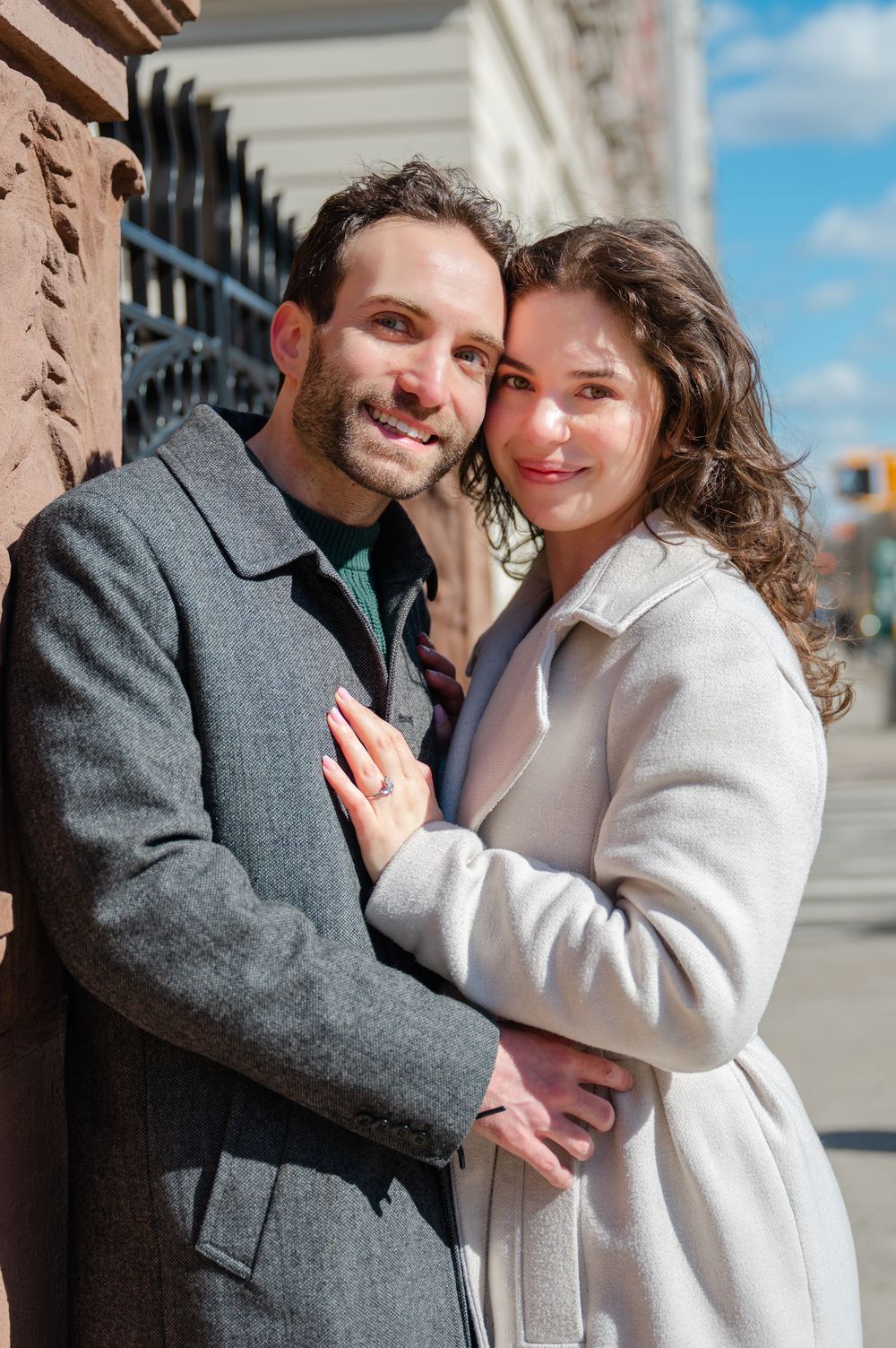 A couple embraces outdoors wearing winter coats, with one in gray and one in cream-colored outerwear on a sunny day.