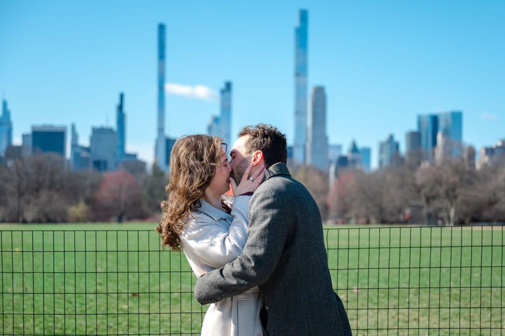 A couple shares a romantic moment in Central Park with the Manhattan skyline visible in the background.