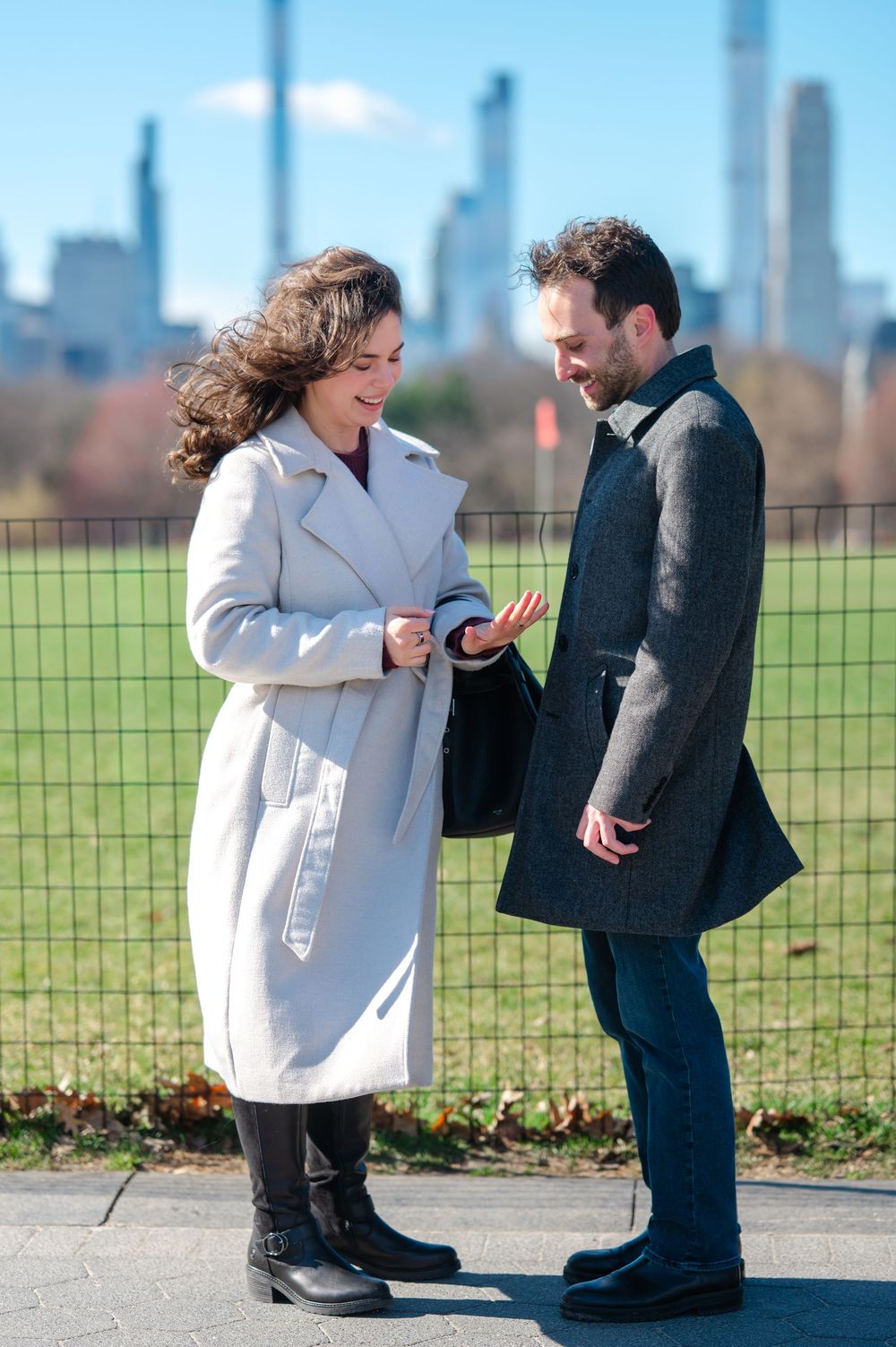 A couple in winter coats stands together on a path with an urban skyline visible in the background on a sunny day.