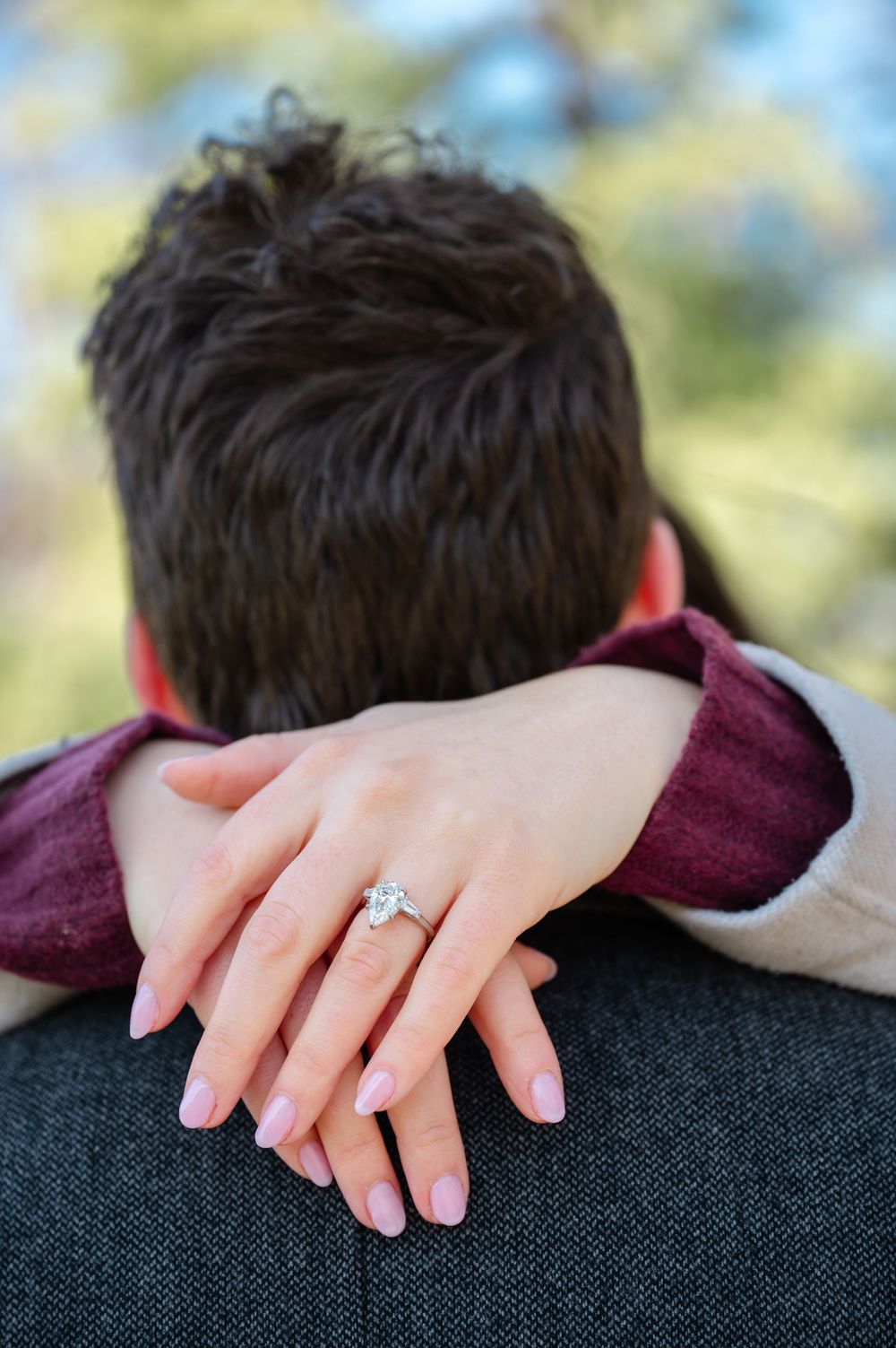 A close-up of hands showing off a diamond engagement ring, with one hand resting on a shoulder in burgundy clothing.