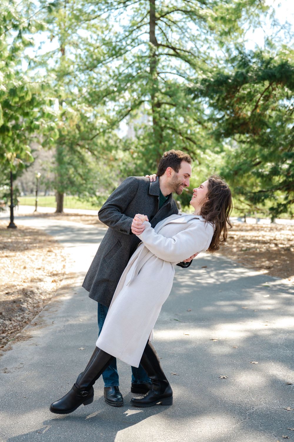 A couple in winter coats share a romantic dance pose on a tree-lined path in a park on a sunny day.