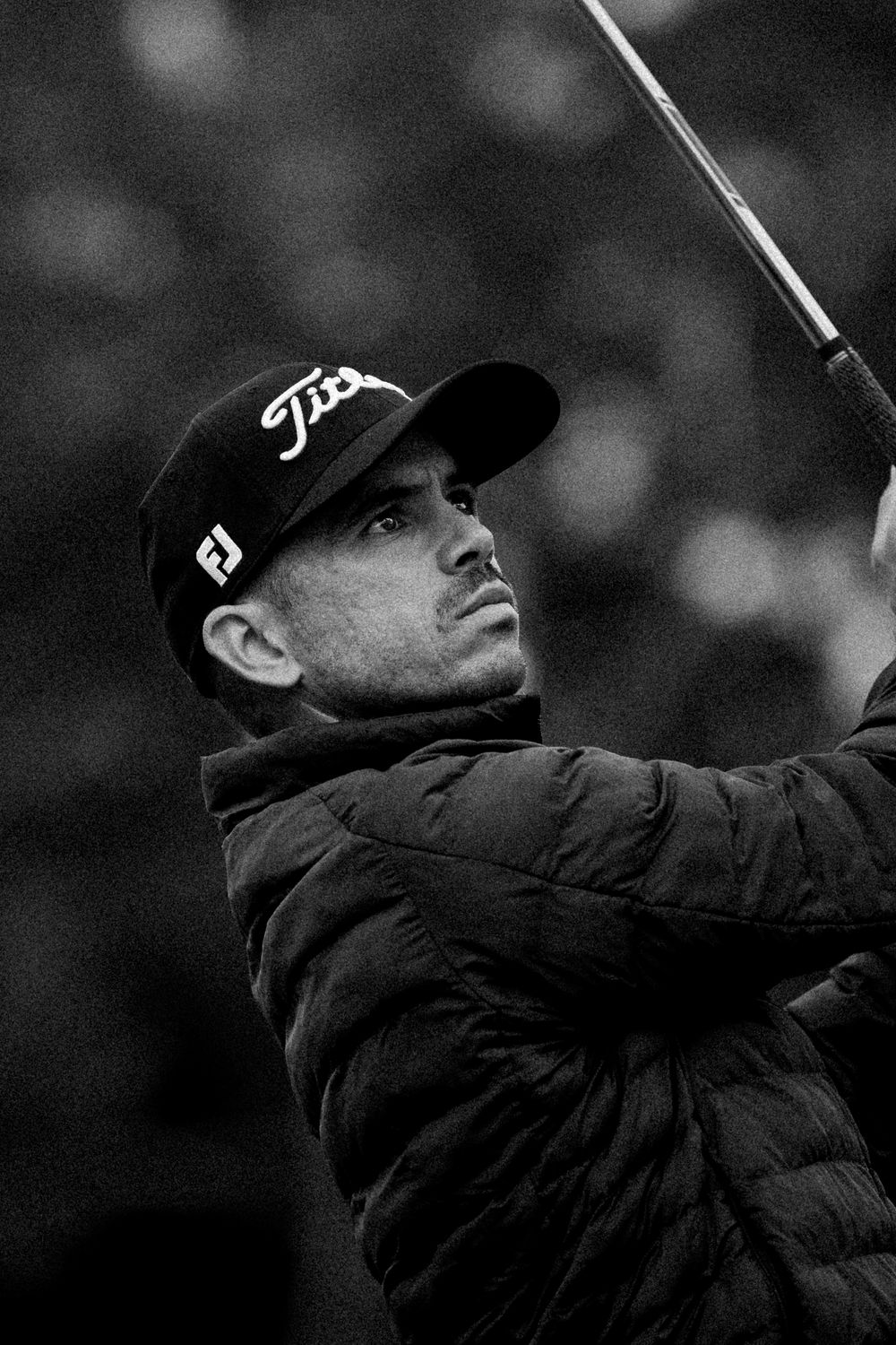 Black and white photo of a golfer wearing a Titleist cap mid-swing, dressed in a puffer jacket on the course.