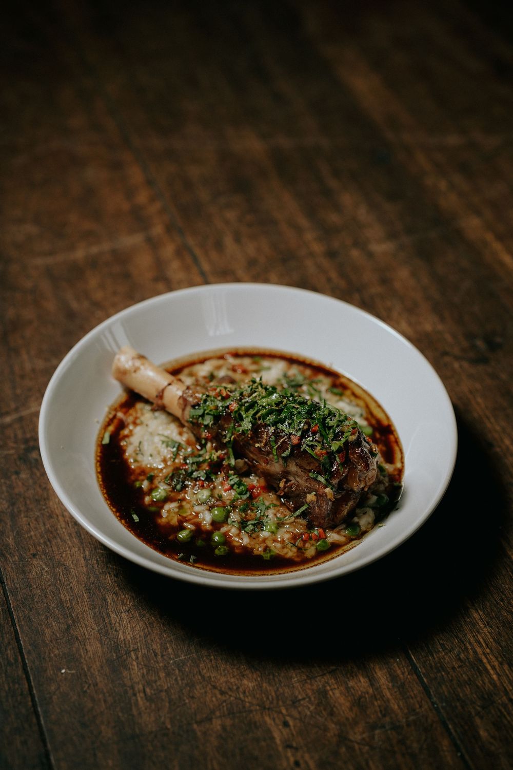 Braised lamb shank in rich red sauce topped with fresh herbs, served in a white bowl on rustic wood table.