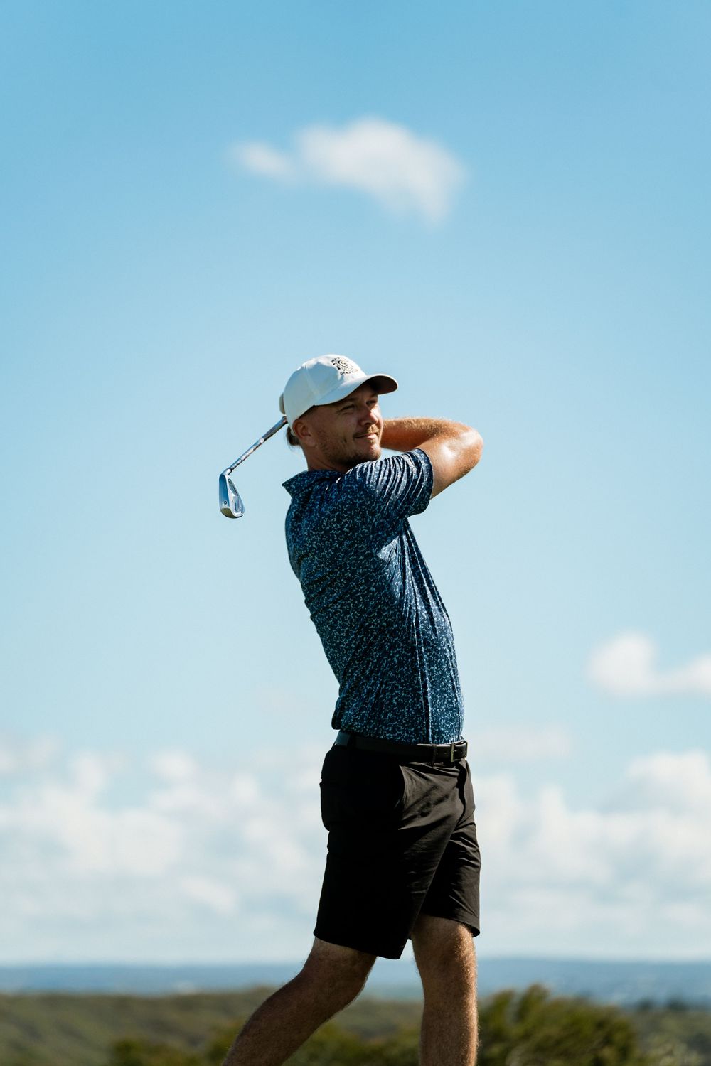 Golfer in navy patterned polo and black shorts swings club against blue sky with white clouds.