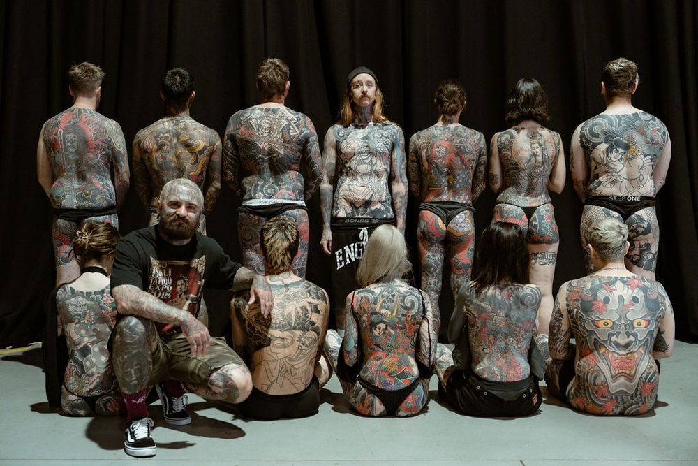 A group of people with intricate tattoo art covering their backs sit in rows on a dark floor.