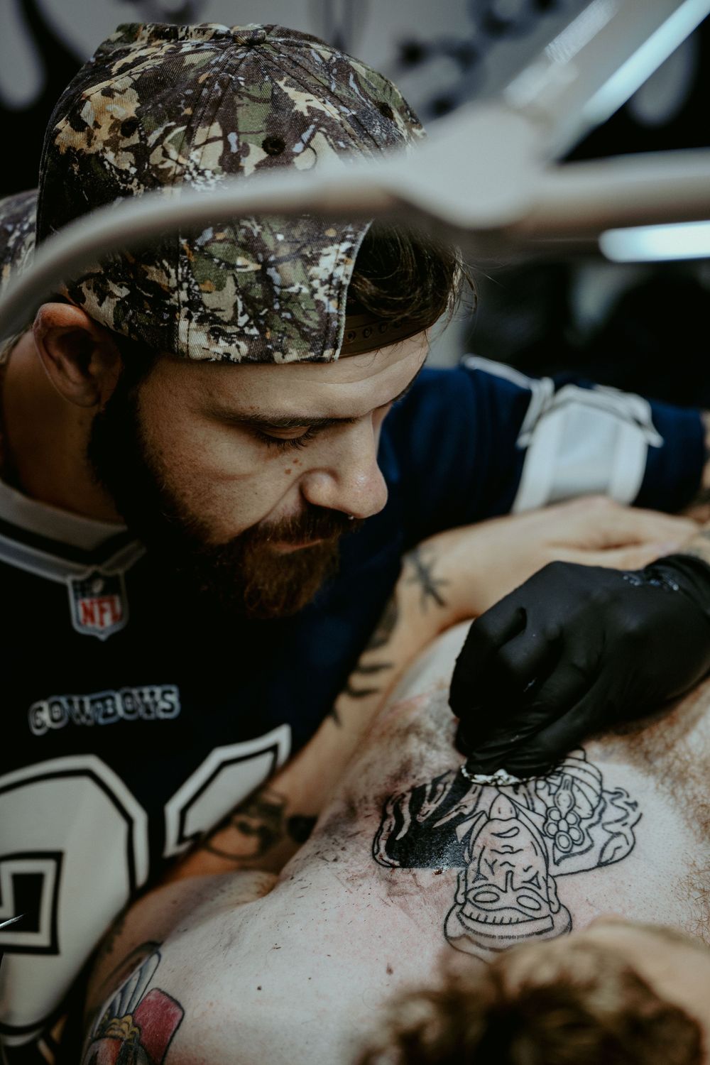 Tattoo artist wearing NFL gear and camo bandana works on detailed tattoo design during professional session.