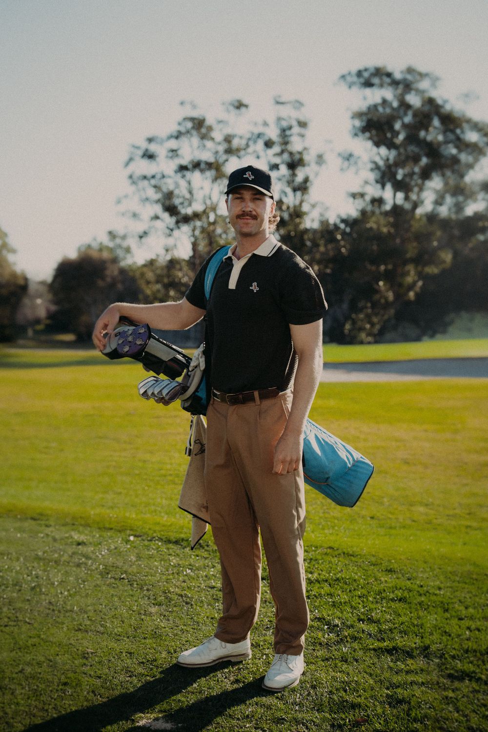 Man in black polo and khakis carrying golf bag on sunny green golf course.