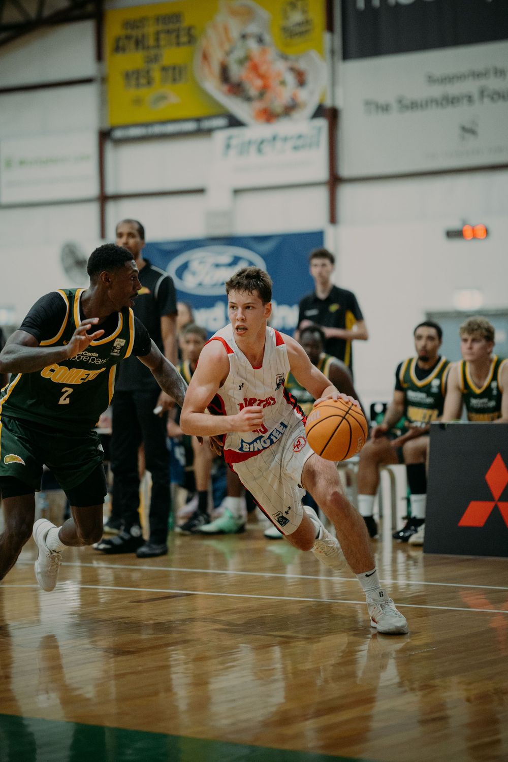 A basketball player in white dribbles past a defender in yellow-green during an indoor game.
