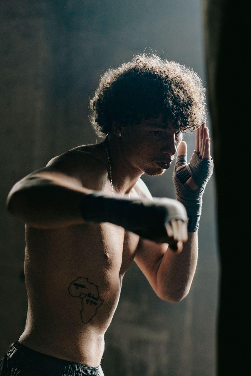 Athletic shirtless boxer with curly hair and Africa tattoo in fighting stance during moody indoor training session.