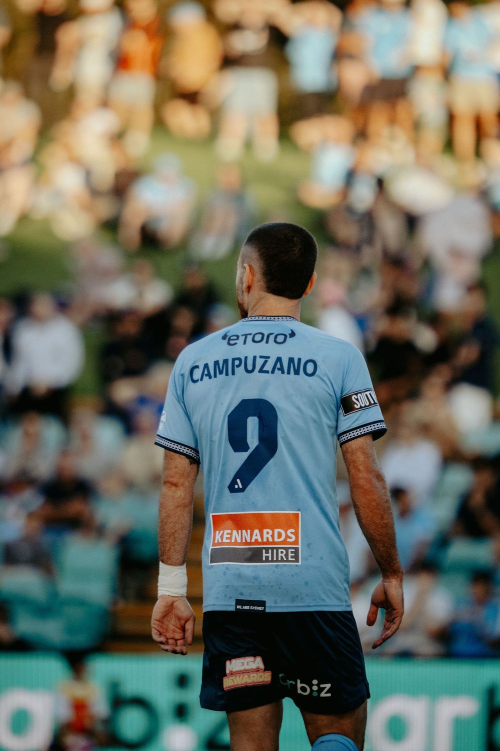 Rugby players in light blue jerseys on a field with spectators visible in the background.