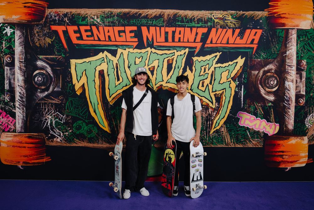 Two people pose with skateboards in front of a colorful Teenage Mutant Ninja Turtles graffiti wall display.