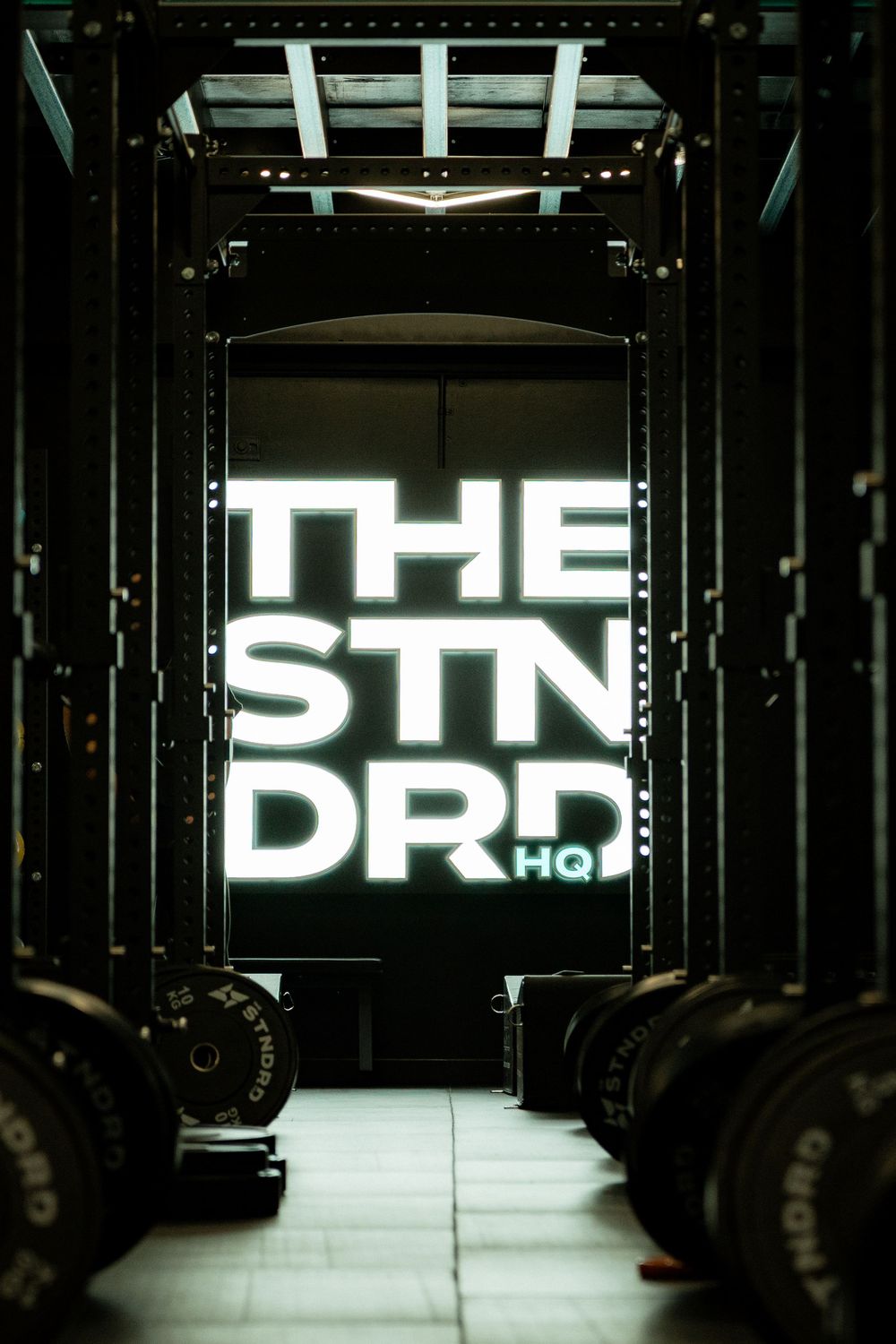 Illuminated neon sign reading 'THE STN DRD HQ' glows between gym equipment in a dark industrial space.
