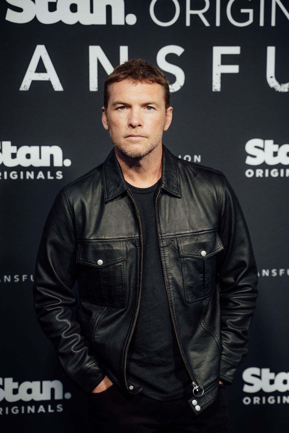 A person in a black leather jacket poses against a dark background with Stan Originals branding.