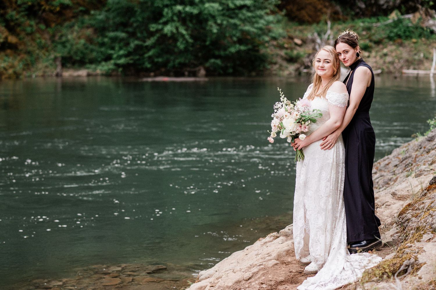 A couple in wedding attire stands on a rocky riverbank surrounded by lush forest and turquoise water.