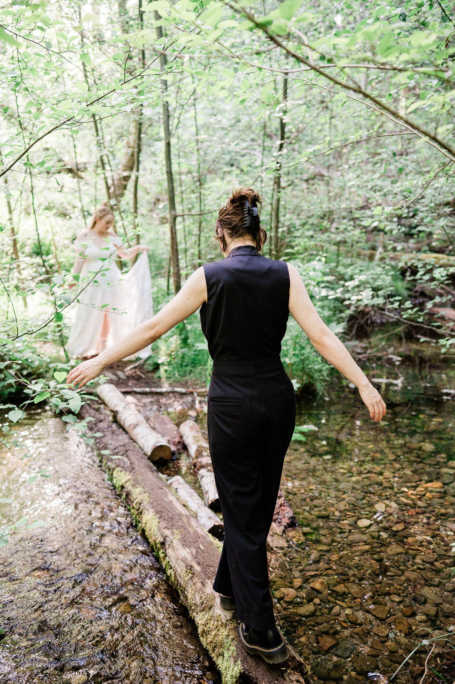 A bride wearing a black pantsuit balances while crossing a fallen log over a shallow forest stream.