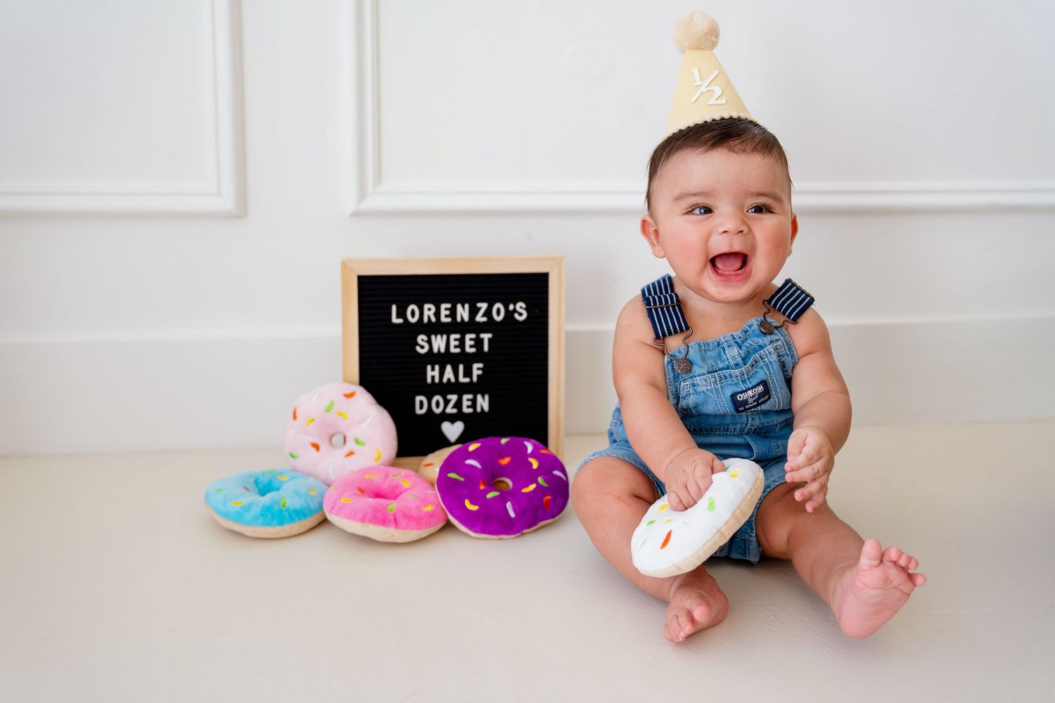 A baby in denim overalls sits on a white floor with colorful donuts and a letter board nearby.