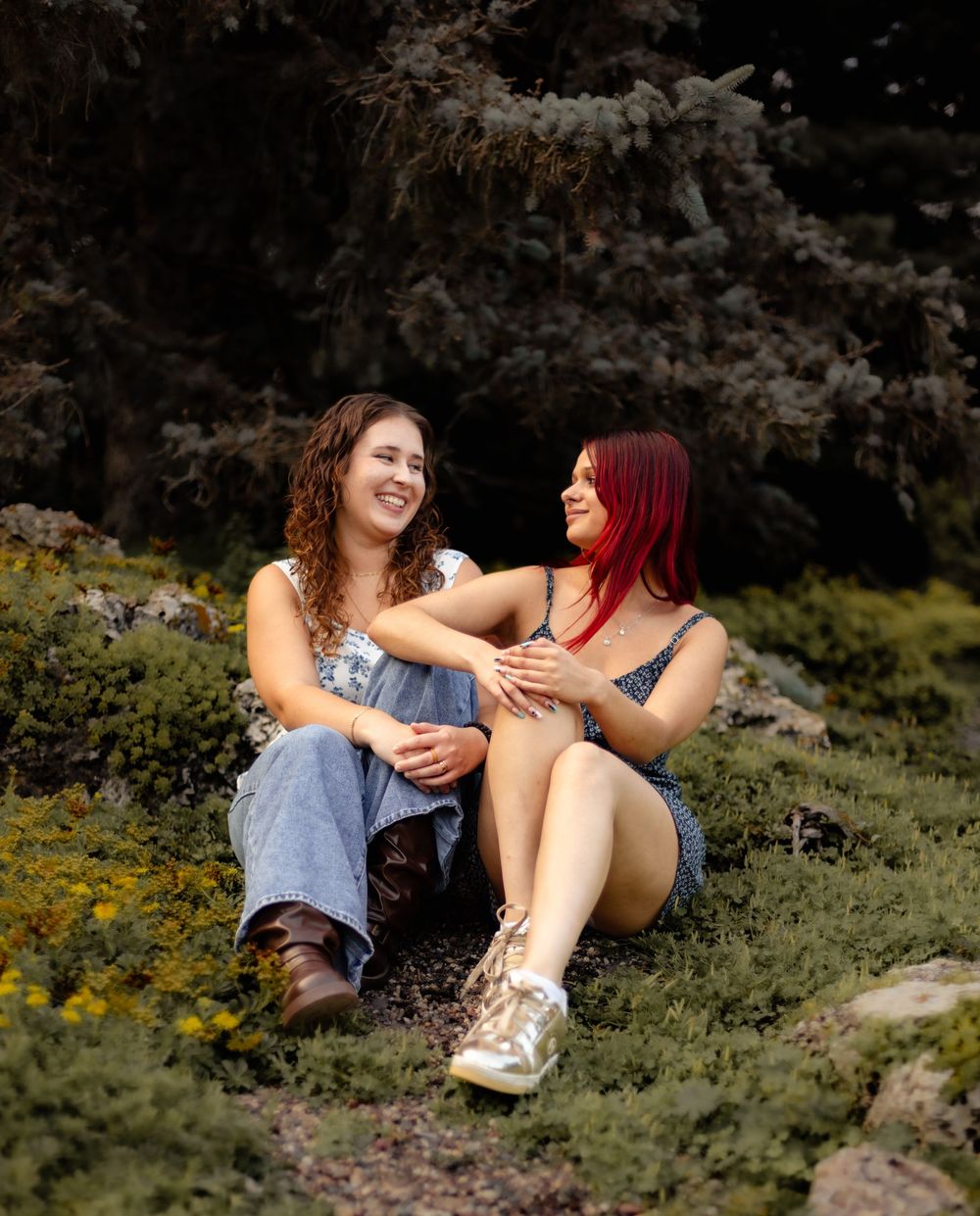 Two friends enjoying a casual moment sitting together outdoors among wildflowers and evergreen trees.
