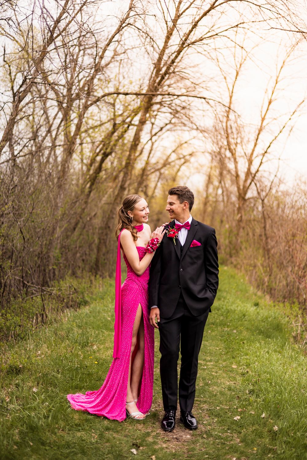 A couple in prom attire, pink gown and black tuxedo, pose on a grassy path surrounded by trees.