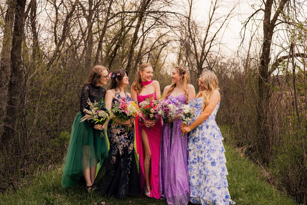 Five women in colorful formal gowns holding bouquets pose together outdoors among bare trees.