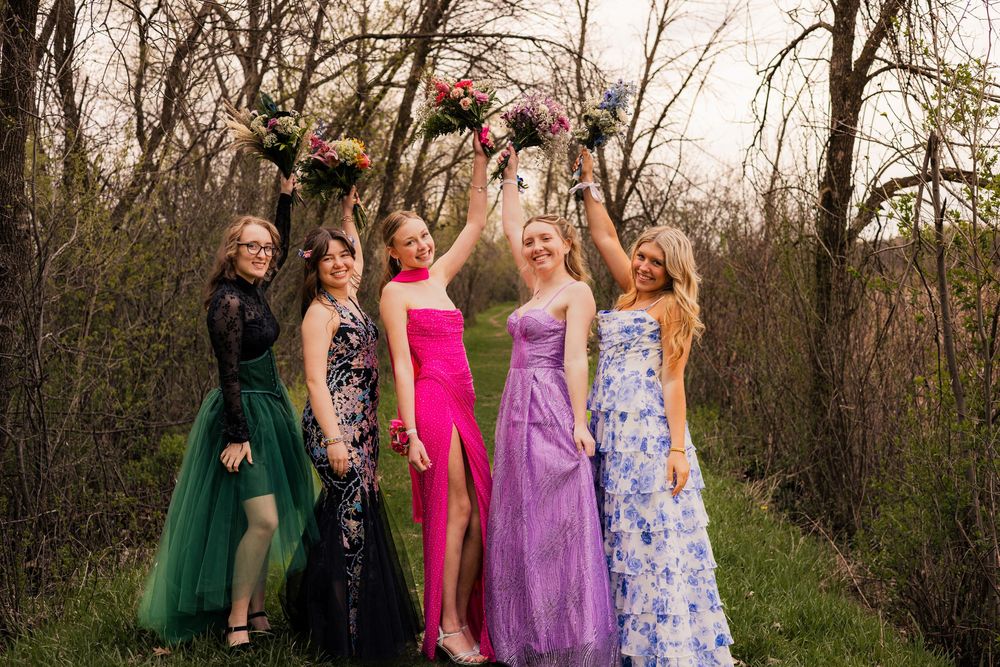 Five young women in colorful formal gowns holding bouquets outdoors among bare trees, dressed for prom.