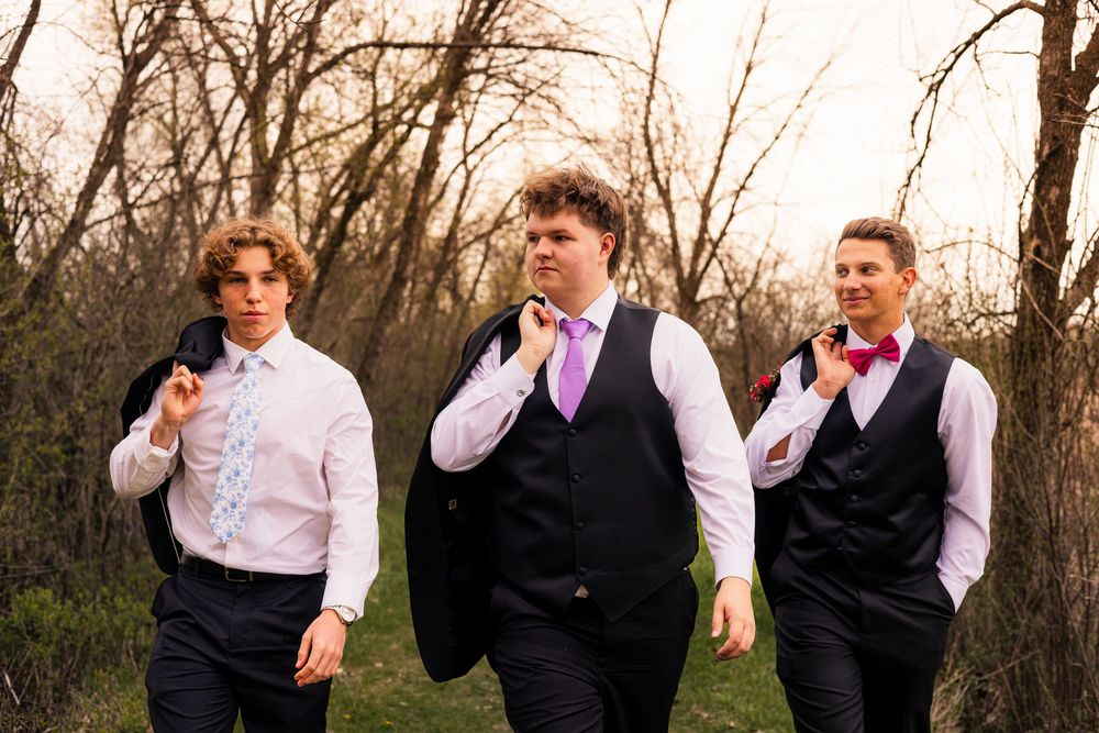 Three young men in formal attire with ties and vests walk outdoors among bare trees, dressed for prom.