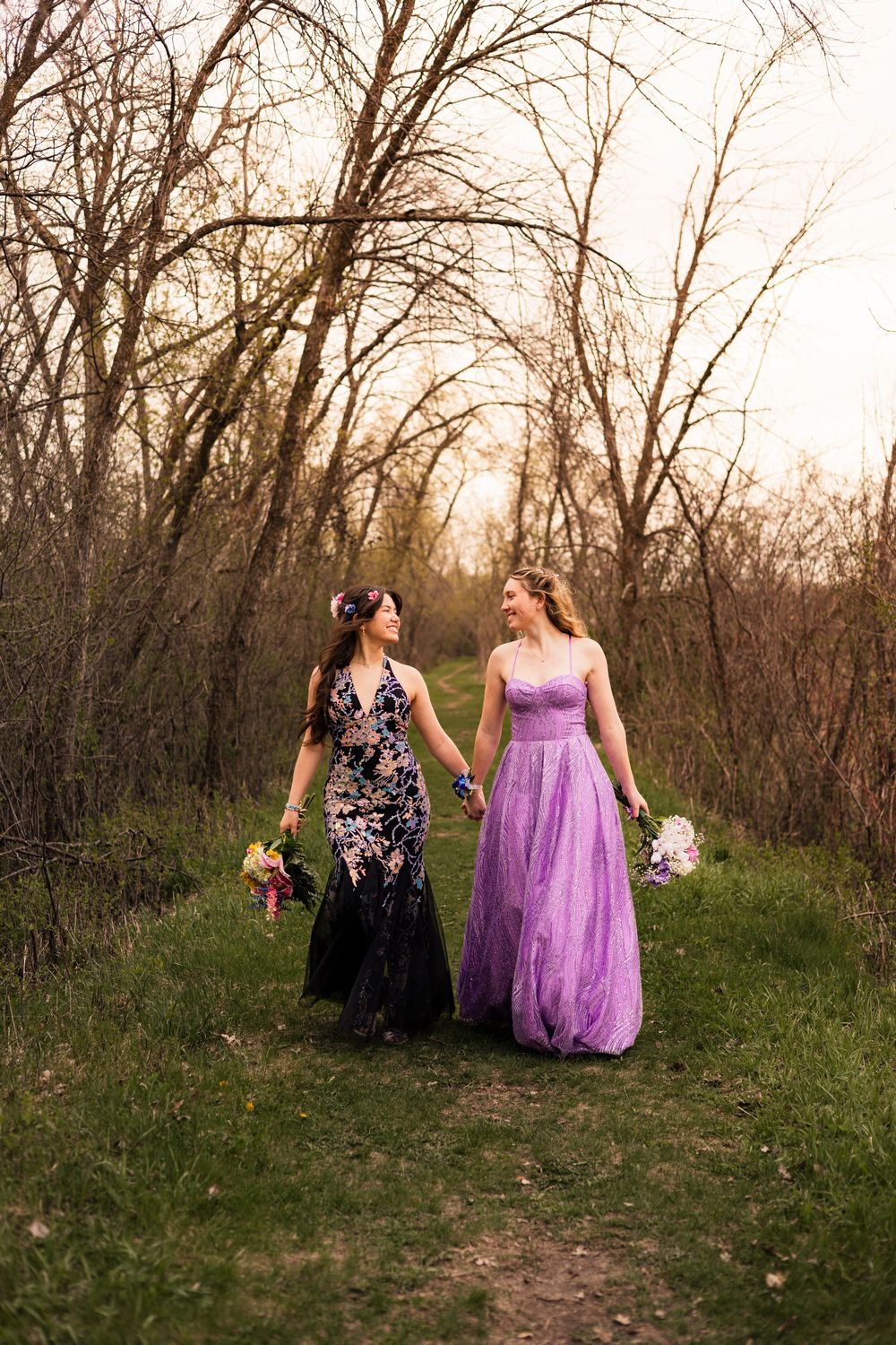 Two women in formal gowns hold hands walking through a wooded path, one in black floral and one in purple tulle.