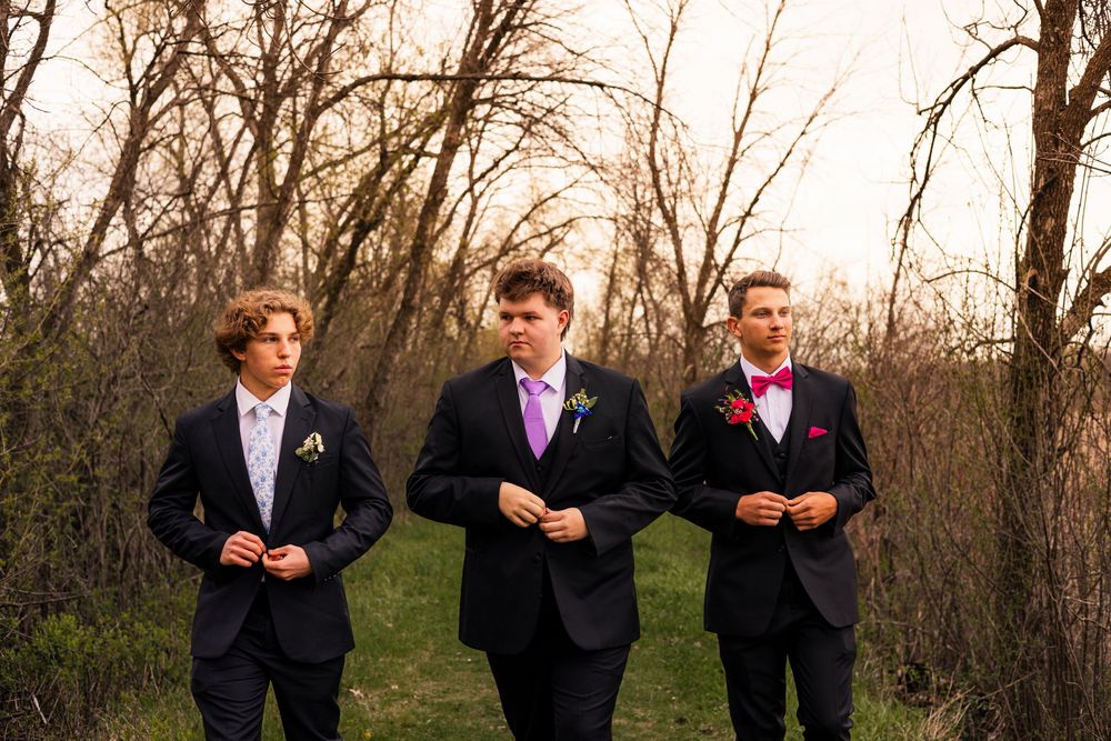 Three young men in dark suits with colorful boutonnieres walk through a wooded area during prom season.