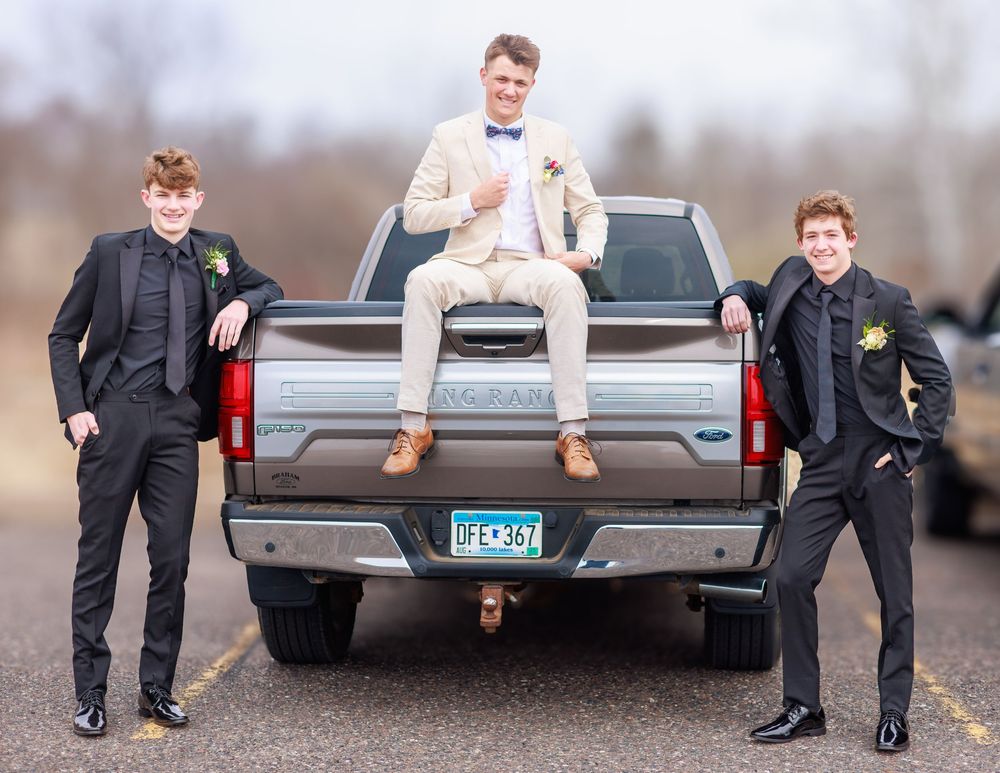 Three young men in formal prom attire pose by a pickup truck tailgate in a parking lot.