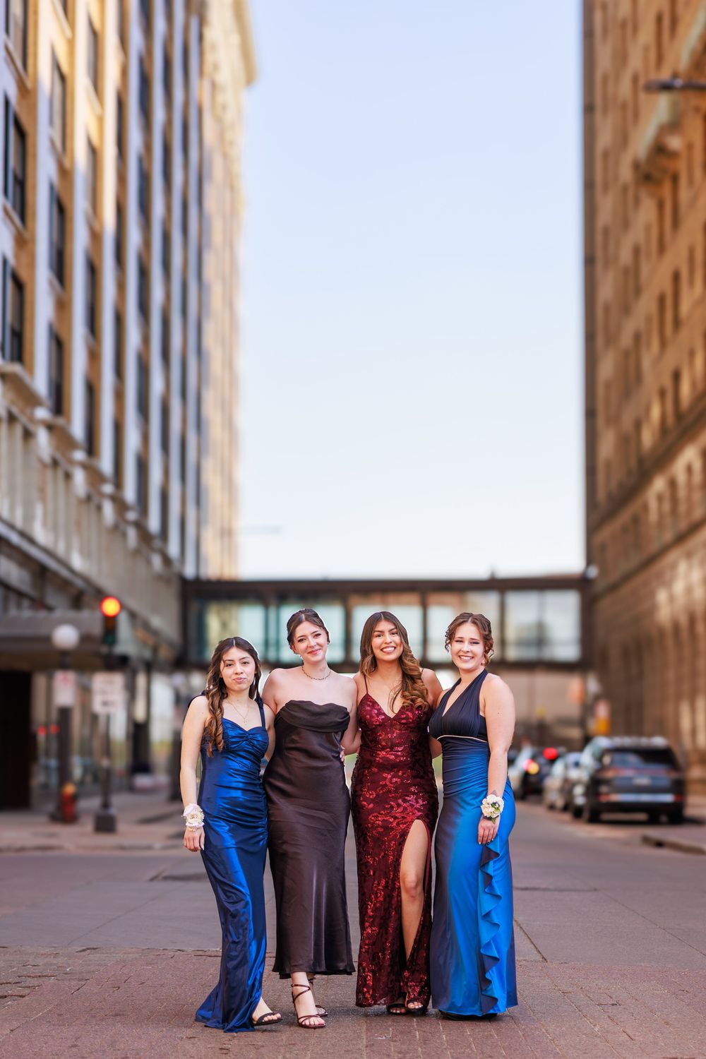 Four young women in elegant prom gowns pose together on a city street at dusk.