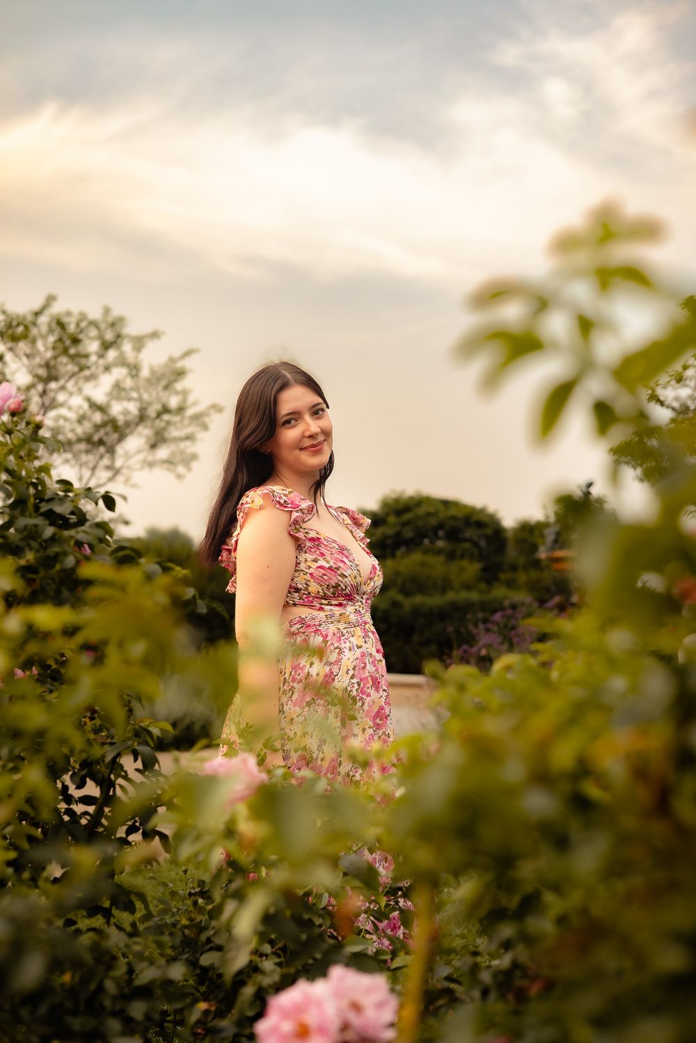 Young woman in floral dress smiling among pink roses in a garden at sunset.