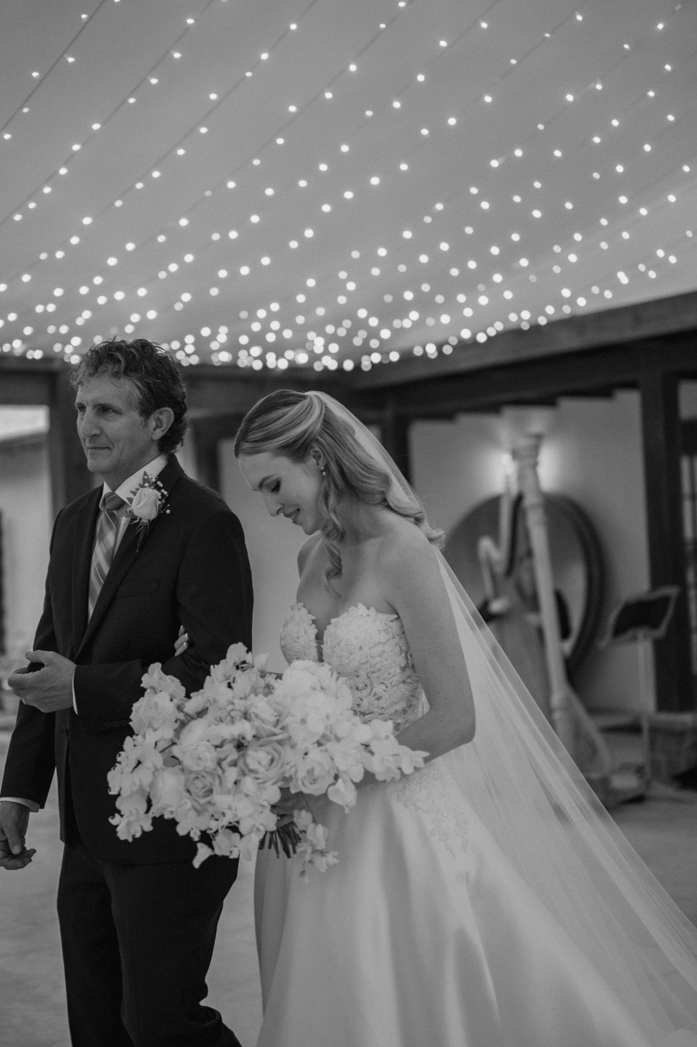 Black and white wedding photo under twinkling string lights with a couple holding a white floral bouquet.