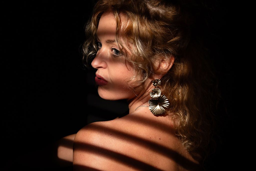 Dramatic portrait with light streaming through venetian blinds creating striped shadows against dark background.