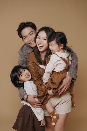 Happy Asian family of four posing together, parents hugging two young daughters, wearing earth tones against beige background.幸福的亞洲一家四口合影